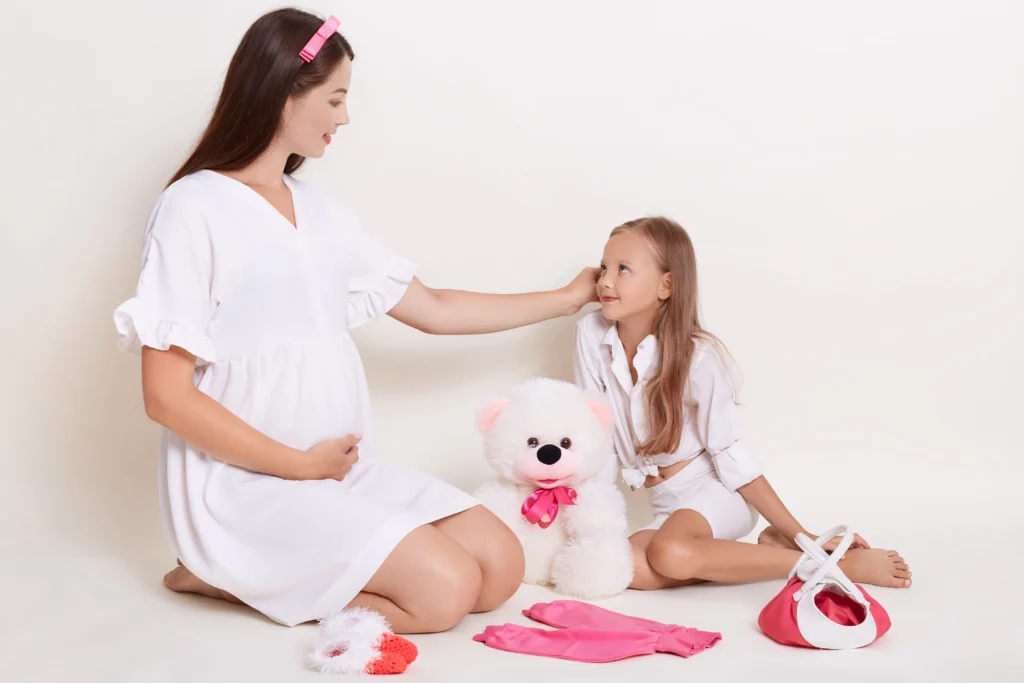 beautiful pregnant woman sitting with doughter floor surrounded with child s clothing soft toy